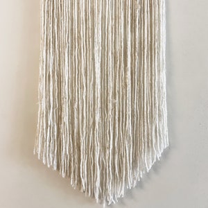 May include: A white macrame wall hanging with a fringe bottom. The hanging is made of yarn and has a natural, bohemian style.