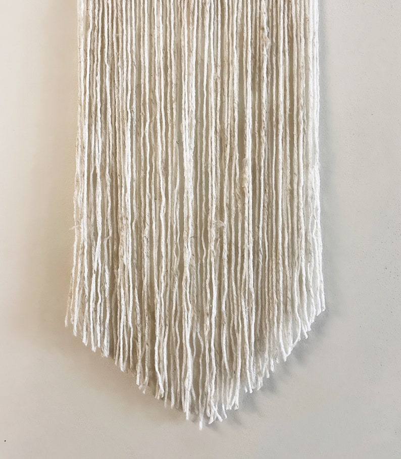May include: A white macrame wall hanging with a fringe bottom. The hanging is made of yarn and has a natural, bohemian style.