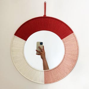 May include: A circular mirror with a rope design in red, cream, and pink. The mirror is suspended on a white wall. The reflection shows a person holding a phone.