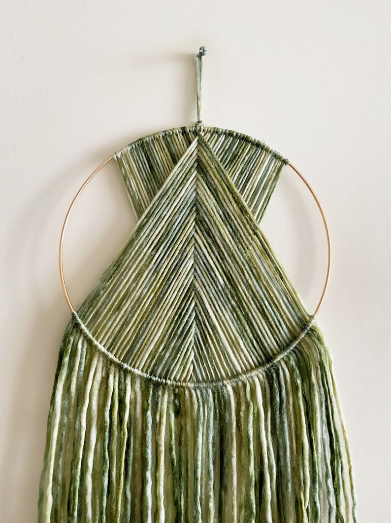 May include: A green and white yarn wall hanging with a geometric design. The hanging is made with a gold metal hoop and has long, flowing strands of yarn.