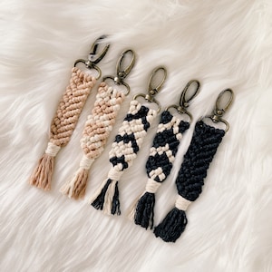 May include: Five macrame keychains in various colors: tan, beige, black and white, and black. Each keychain features a woven design, a tassel, and a bronze-colored clasp for attaching to keys or bags. The keychains are displayed on a white, fluffy surface.