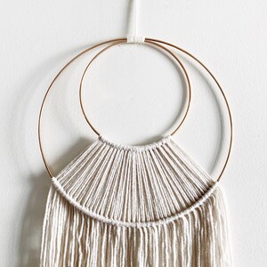 May include: A white macrame wall hanging with a fringe, featuring two concentric gold metal hoops.