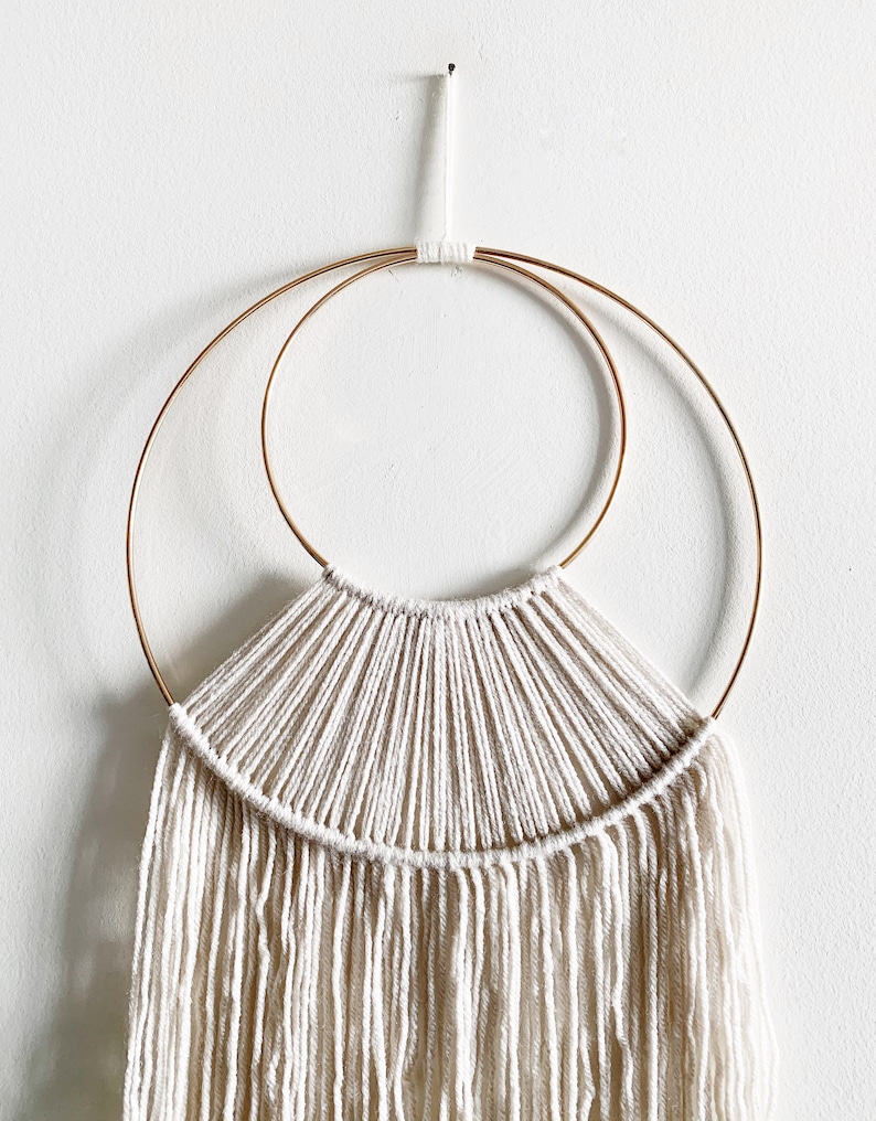May include: A white macrame wall hanging with a fringe, featuring two concentric gold metal hoops.