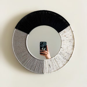 May include: A round mirror with a black and white rope frame. The mirror is reflecting a person holding a smartphone.