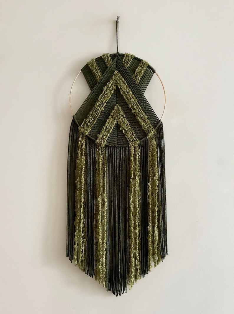May include: A green and gold macrame wall hanging with a geometric design. The hanging features a circular hoop with fringe tassels.