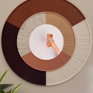May include: A circular wall mirror with a woven design. The frame is divided into sections of brown, tan, and cream colours. The mirror reflects a person holding a phone. A decorative home decor item.