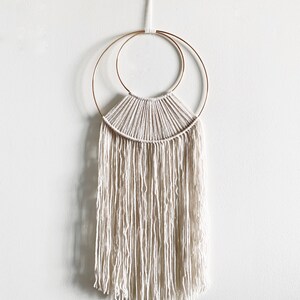 May include: A white macrame wall hanging with a gold metal hoop and a fringe border.