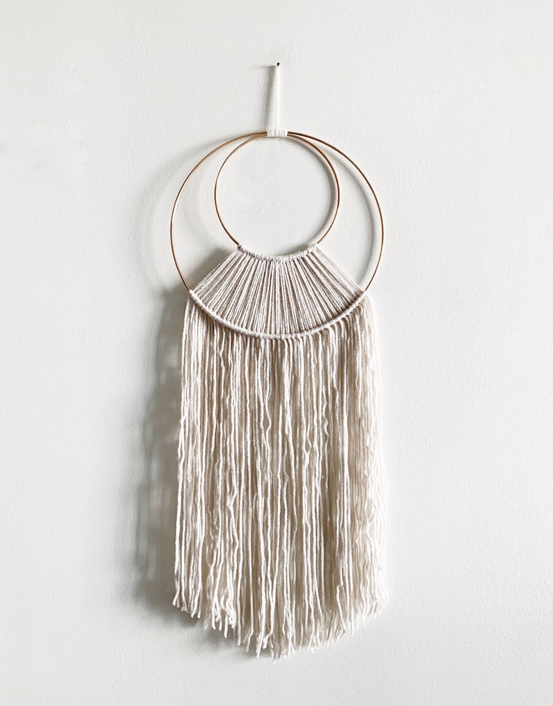 May include: A white macrame wall hanging with a gold metal hoop and a fringe border.