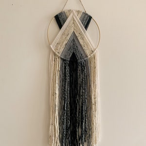 May include: A black, white and grey yarn wall hanging with a gold hoop. The yarn is woven into a geometric pattern and hangs in long strands.