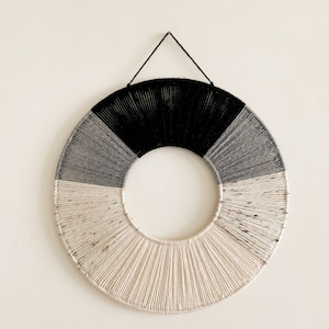 May include: A circular wall hanging made of woven yarn in shades of white, grey, and black. The hanging is attached to the wall with a black string.