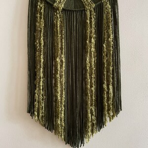 May include: A green and olive green macrame wall hanging with a fringe border. The fringe is made of different textures of yarn.