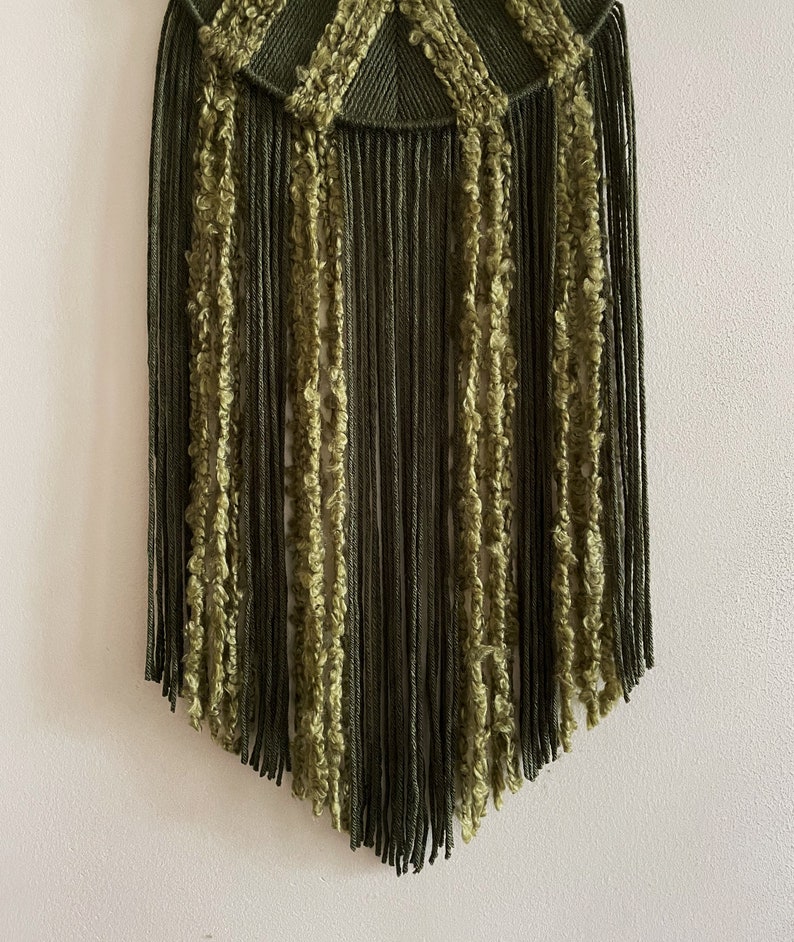 May include: A green and olive green macrame wall hanging with a fringe border. The fringe is made of different textures of yarn.
