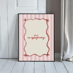 May include: Framed art print with a cream background and pink vertical stripes. The artwork has a red, wavy border and the text "brb... i'm hyperfixating" in red script. The frame is black.