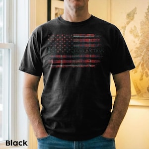 May include: Black t-shirt with a distressed American flag design in red, white, and blue. The flag incorporates a plaid pattern and the text "CLAN RATTRAY". The shirt is short-sleeved.