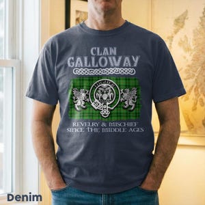 May include: Denim blue t-shirt featuring the text "Clan Galloway" above a green and black plaid design with a crest and the words "Revelry & Mischief Since The Middle Ages". The shirt has short sleeves.