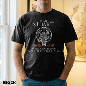May include: A black short-sleeved t-shirt featuring the text CLAN STUART at the top, a circular crest with a lion emblem and the Latin phrase NOBILIS EST IRA LEONIS, and the text WREAKING HAVOC SINCE THE MIDDLE AGES with red thistle accents.