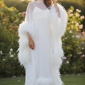 May include: A long, flowing white dress with a sheer overlay and feather trim. The dress features a high neckline and is worn outdoors in a garden setting. The sleeves and hemline are adorned with fluffy white feathers.