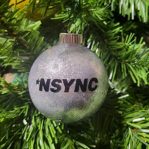 May include: A silver glitter Christmas ornament with the word "NSYNC" printed in black letters.