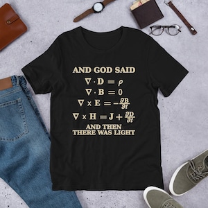 May include: Black t-shirt with the text "AND GOD SAID" above a series of mathematical equations, followed by "AND THEN THERE WAS LIGHT" in a light beige color. The shirt is displayed on a light gray surface.