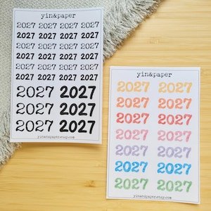 Puede incluir: Dos hojas de pegatinas rectangulares con el año 2027 impreso en varias fuentes y colores. La hoja de la izquierda muestra el texto 2027 en negro, mientras que la de la derecha presenta filas de 2027 en amarillo, naranja, rojo, rosa, morado, azul y verde.
