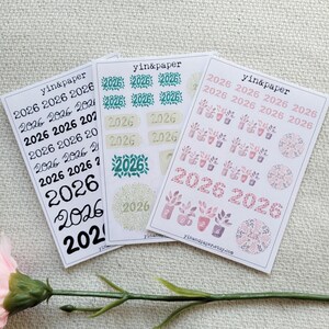 Op de afbeelding: Drie vellen stickers met het jaar 2026 in verschillende lettertypen en ontwerpen. De stickers bevatten bloempatronen en zijn verkrijgbaar in zwart, groen en roze. De merknaam "yin&paper" staat op elk vel.
