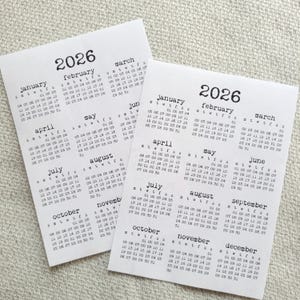 May include: Two small, white 2026 calendars with a simple, minimalist design. Each calendar displays the months of the year with a clean, sans-serif font. The calendars are arranged on a neutral surface.