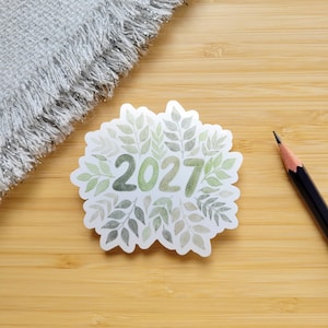 Könnte beinhalten: Ein gestanzter Aufkleber mit der Jahreszahl 2027 in einer fetten, abgerundeten Schrift, umgeben von einer Aquarell-Illustration aus verschiedenen grünen Blättern und Pflanzen. Der Aufkleber hat einen weißen Rand auf einer hellen Holzoberfläche.