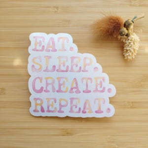 May include: A white sticker with the words "EAT. SLEEP. CREATE. REPEAT." in pink and yellow watercolor lettering. The sticker is on a light brown wooden surface, with dried flowers in the upper right corner.