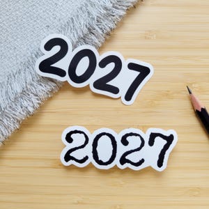 Op de afbeelding: Twee gestanste stickers met het jaartal 2027 in zwarte letters. De bovenste sticker toont de cijfers in een glad, vet lettertype, terwijl de onderste sticker dezelfde cijfers weergeeft in een grillige, ruwe of handgetekende stijl.