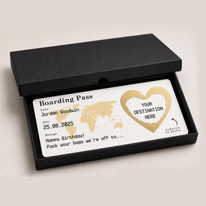 May include: A white boarding pass with gold accents, presented in a black box. The pass includes a world map, a heart-shaped scratch-off area labeled "Your Destination Here," and the text "Happy Birthday! Pack your bags we're off to..."