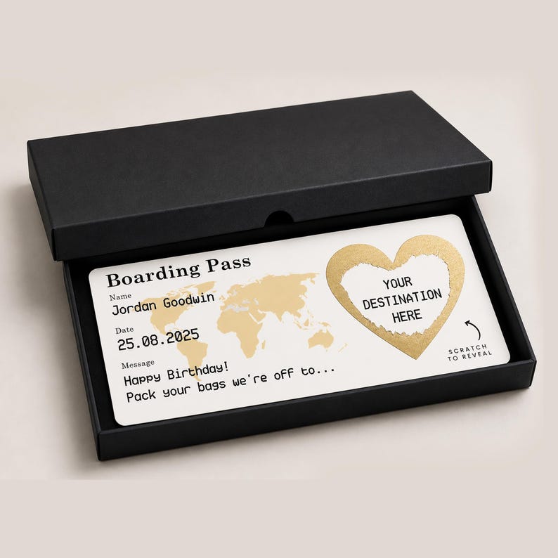 May include: A white boarding pass with gold accents, presented in a black box. The pass includes a world map, a heart-shaped scratch-off area labeled "Your Destination Here," and the text "Happy Birthday! Pack your bags we're off to..."