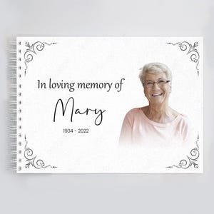 May include: A white memorial book with a black spiral binding. The book has a black and white floral design on the cover and the text "In loving memory of Mary 1934 - 2022". A photo of a woman is in the top right corner of the book.