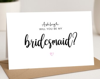 May include: A white card with black text that reads "Ashleigh, will you be my bridesmaid?" with a pink heart below the text.