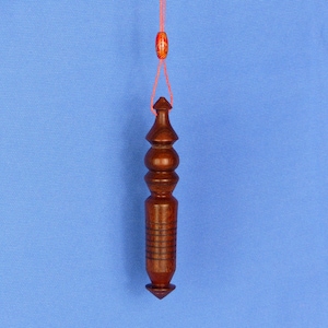 Peut inclure: Un pendule en bois brun foncé, avec une base pointue et une série de sections arrondies. Il est suspendu à une ficelle rouge avec une petite perle décorative. Le fond est bleu uni.
