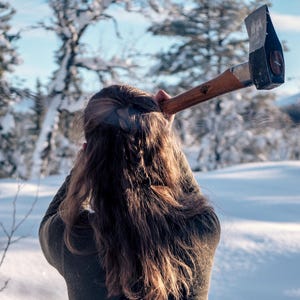 May include: A person swinging an axe in a snowy forest. The axe has a wooden handle and a metal head. The person is wearing a green sweater and black overalls. A tree stump with meat on it is in the foreground.