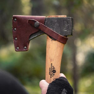 May include: Axe with a wooden handle and a dark metal head, protected by a brown leather sheath. The handle has the text "Gränsfors Bruk Sweden" printed on it. The axe is held in a gloved hand against a blurred green background.