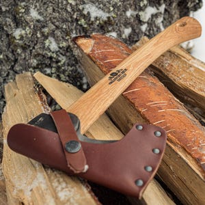 Può includere: Un'ascia piccola con manico in legno e testa in metallo scuro, appoggiata su un mucchio di legna da ardere. L'ascia ha un fodero in pelle marrone. Il manico riporta la scritta "Gransfors Bruk Sweden".