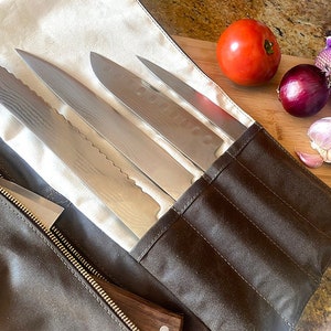 Könnte beinhalten: Eine braune Leder-Messerrolle mit vier Fächern für Messer. Die Rolle ist geöffnet und vier silberne Messer sind sichtbar. Die Rolle liegt auf einem Holzbrett mit einer roten Tomate, einer violetten Zwiebel und drei Knoblauchzehen.