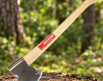 Può includere: Un'accetta con testa in metallo lucido e manico in legno chiaro. Un adesivo rettangolare rosso è applicato sul manico, con la scritta bianca HACHA ARTESANAL e un piccolo logo. L'accetta è appoggiata su un ceppo d'albero.