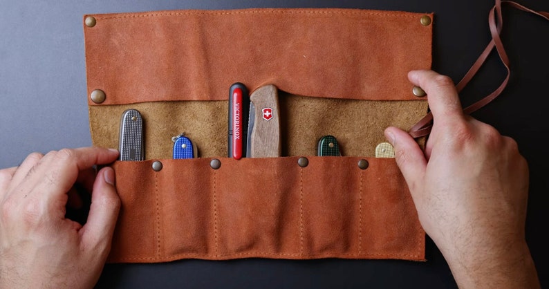 May include: A brown leather roll with six slots for holding pocket knives. The roll is open and has five knives inside, including a red and black Victorinox knife.