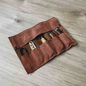 May include: A brown leather roll-up case with six slots for holding tools. The case is rolled up and secured with a flap. The case is made of a soft, supple leather and has a rustic look.