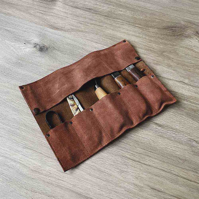 May include: A brown leather roll-up case with six slots for holding tools. The case is rolled up and secured with a flap. The case is made of a soft, supple leather and has a rustic look.