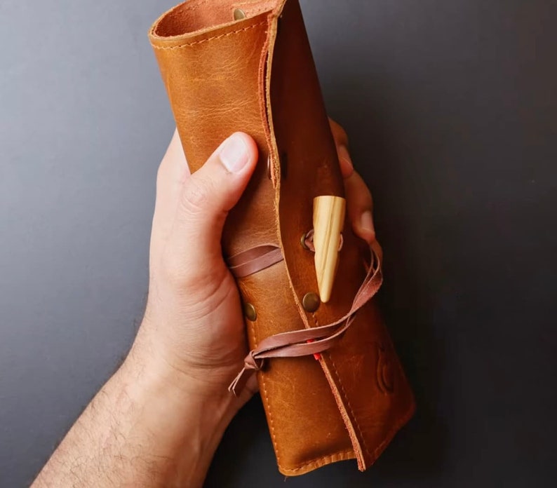 May include: A brown leather roll-up case with a wooden dowel attached. The case is held in a person's hand.