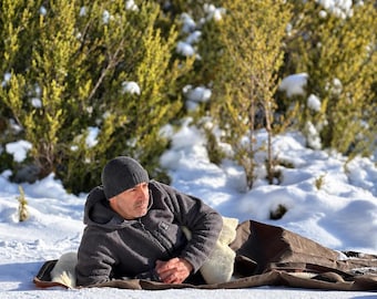May include: A charcoal gray fleece zip-up hoodie with a matching knit beanie, worn while lying on a brown, fur-lined blanket outdoors in a snowy environment with green, snow-dusted bushes in the background.