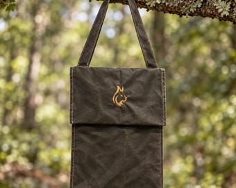 Può includere: Una borsa a tracolla in tela cerata verde oliva scuro con patta ripiegabile e un unico cinturino. La parte anteriore della borsa presenta un piccolo logo ricamato a forma di testa di volpe giallo oro, centrato sulla patta superiore.