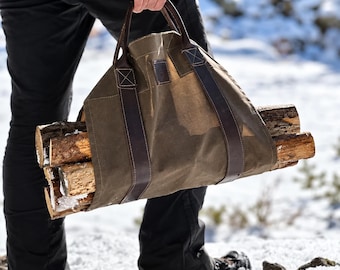 May include: An olive green waxed canvas firewood carrier featuring dark brown leather handles and reinforced stitching. The carrier is filled with several pieces of rough-cut wooden logs, showcasing a durable design for transporting firewood.