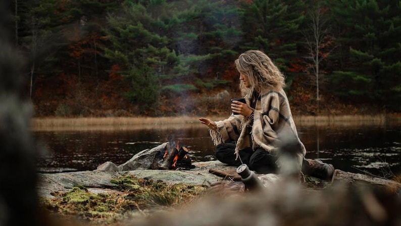 May include: A woman sits by a small campfire on a rocky lakeshore, wearing a brown and white patterned poncho and holding a mug. The trees in the background are bare and the water is still.
