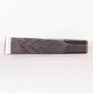 Pode incluir: Uma cunha de metal cinza escuro com um padrão de chevrão na parte superior. A cunha tem uma borda plana e brilhante de cor prateada e uma extremidade facetada. Projetada para rachar madeira.