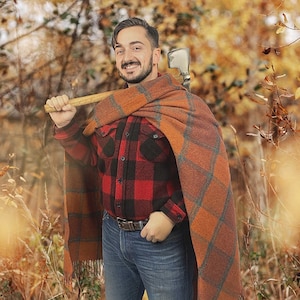 May include: A man wearing a red and black plaid shirt, blue jeans, and a brown and orange plaid blanket. He is holding a wooden-handled axe over his shoulder. The background is a blurred image of fall foliage.