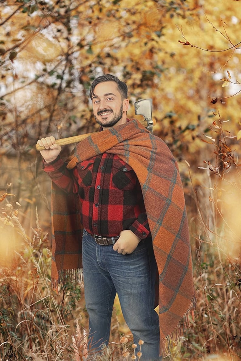May include: A man wearing a red and black plaid shirt, blue jeans, and a brown and orange plaid blanket. He is holding a wooden-handled axe over his shoulder. The background is a blurred image of fall foliage.